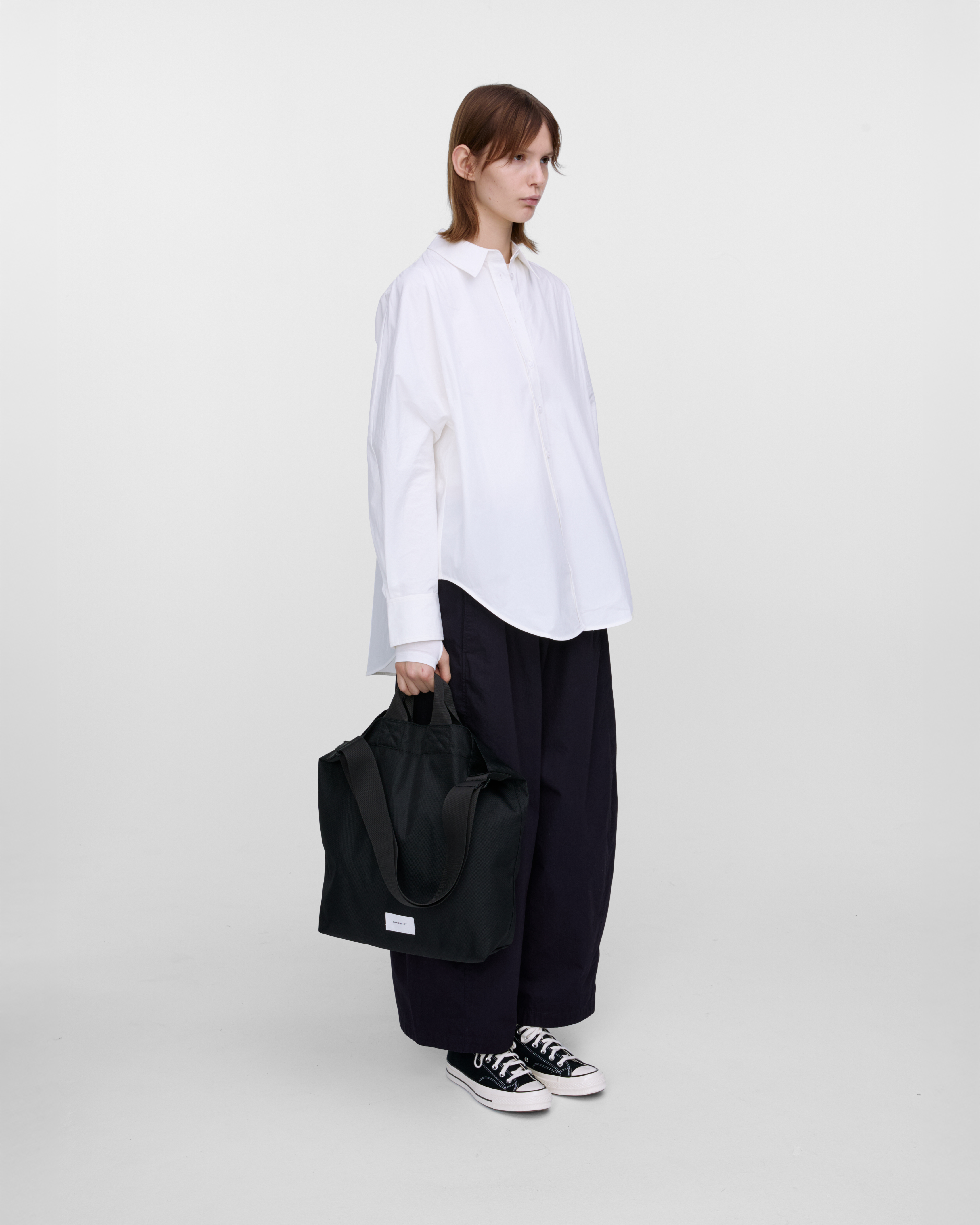 Female model holding Sandqvist Ground Shopper Bag, black. Side view, holding the bag in one hand.
