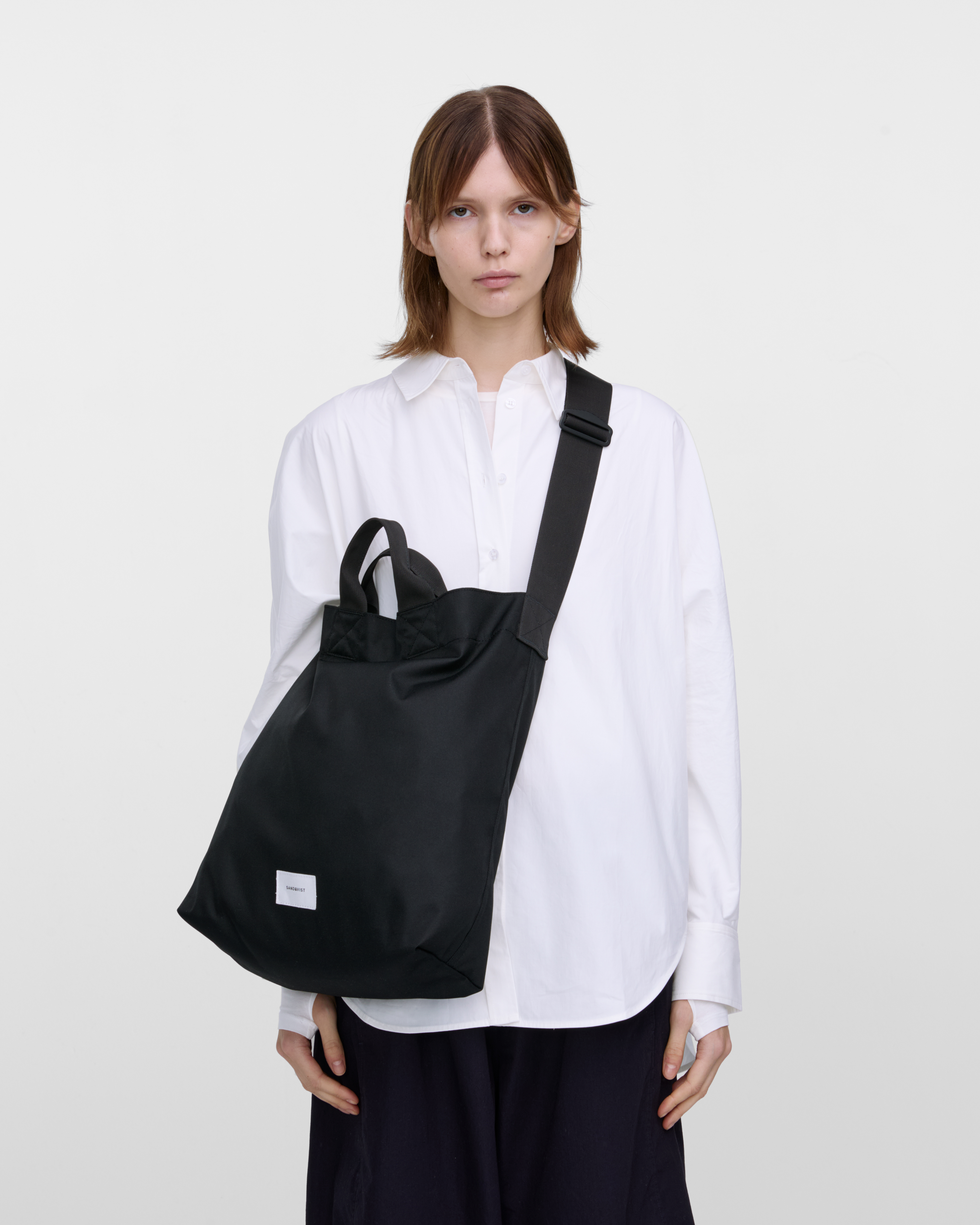 Female model wearing Sandqvist Ground Shopper Bag, black. Front view, wearing the bag on one shoulder crossbody.