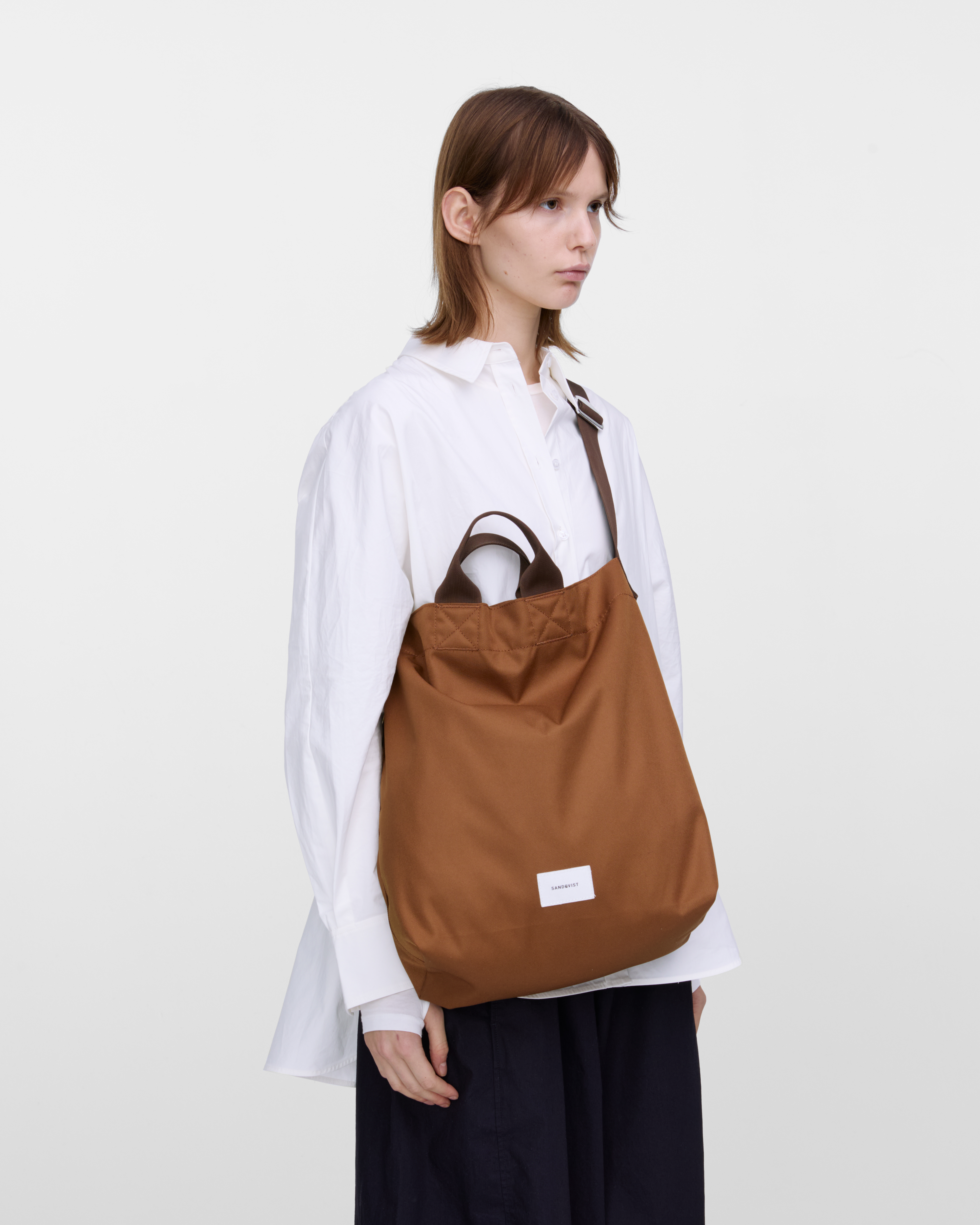 Mannequin féminin portant le sac Sandqvist Ground Shopper Bag, marron. Vue de profil, portant le sac en bandoulière sur une épaule.
