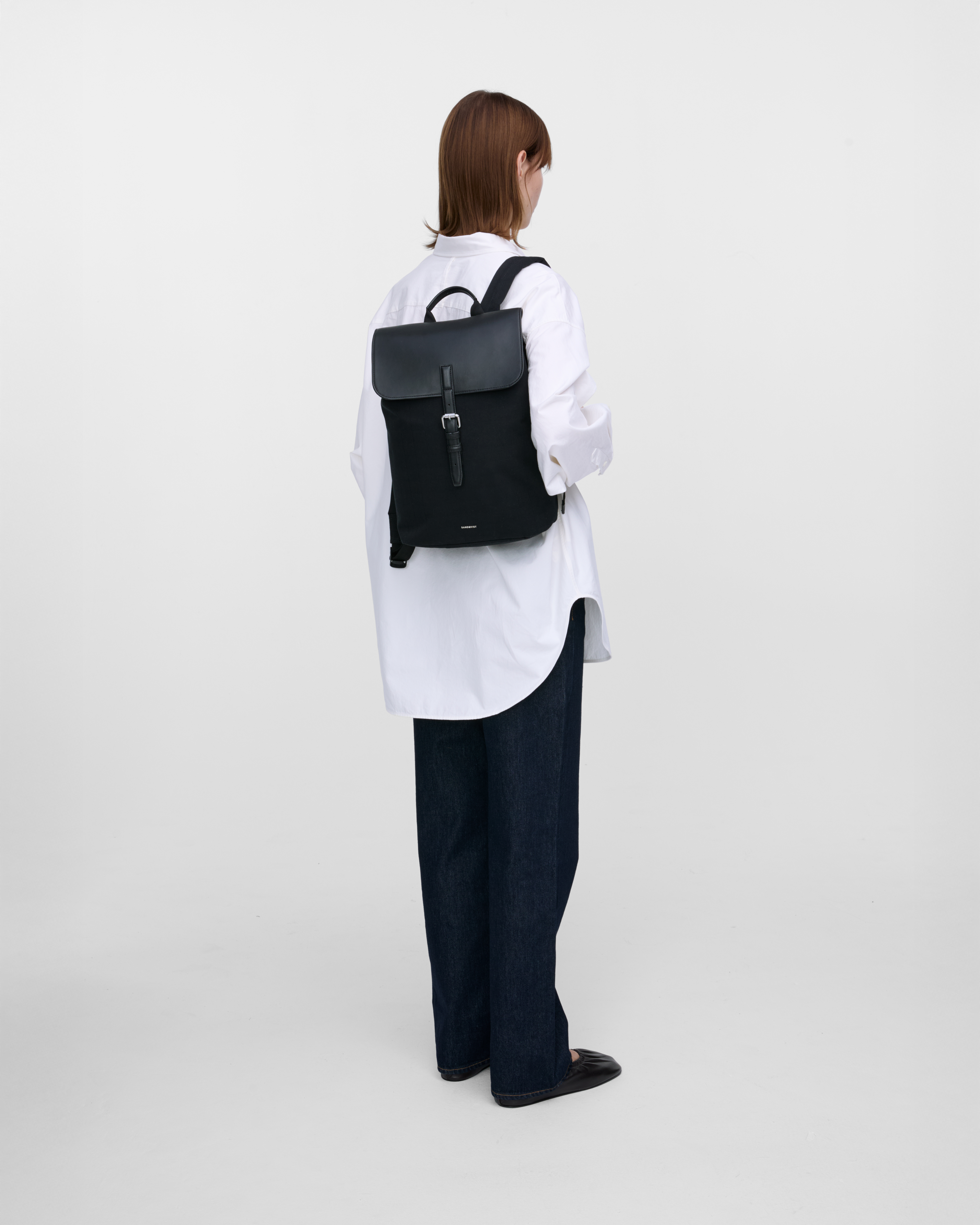 Female model wearing Sandqvist Everyday Lid Backpack M, black. Back view, wearing the bag on one shoulder with the bag in the back.
