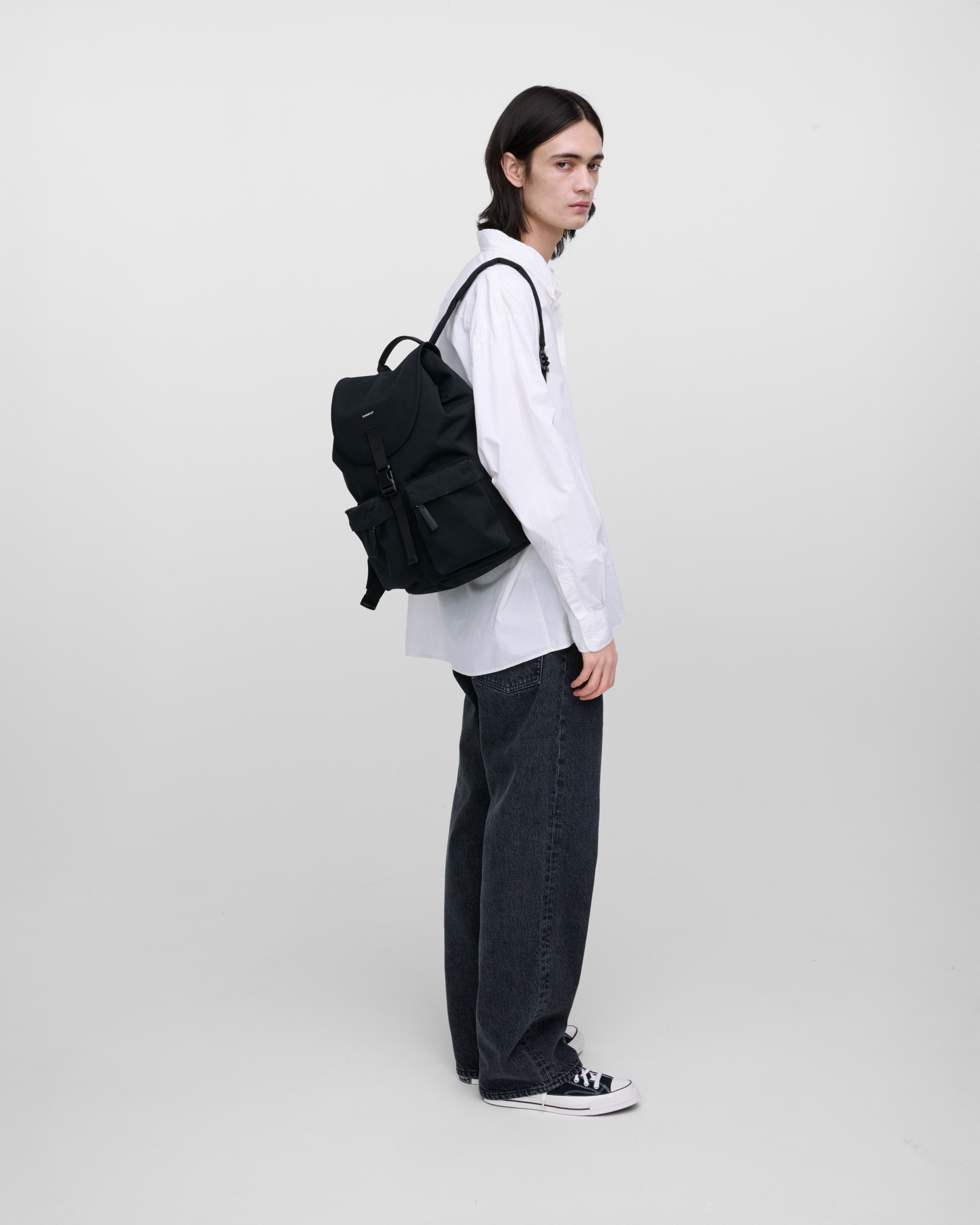 Male model wearing Sandqvist Everyday Rucksack, black. Side view, wearing the bag on one shoulder on the back.