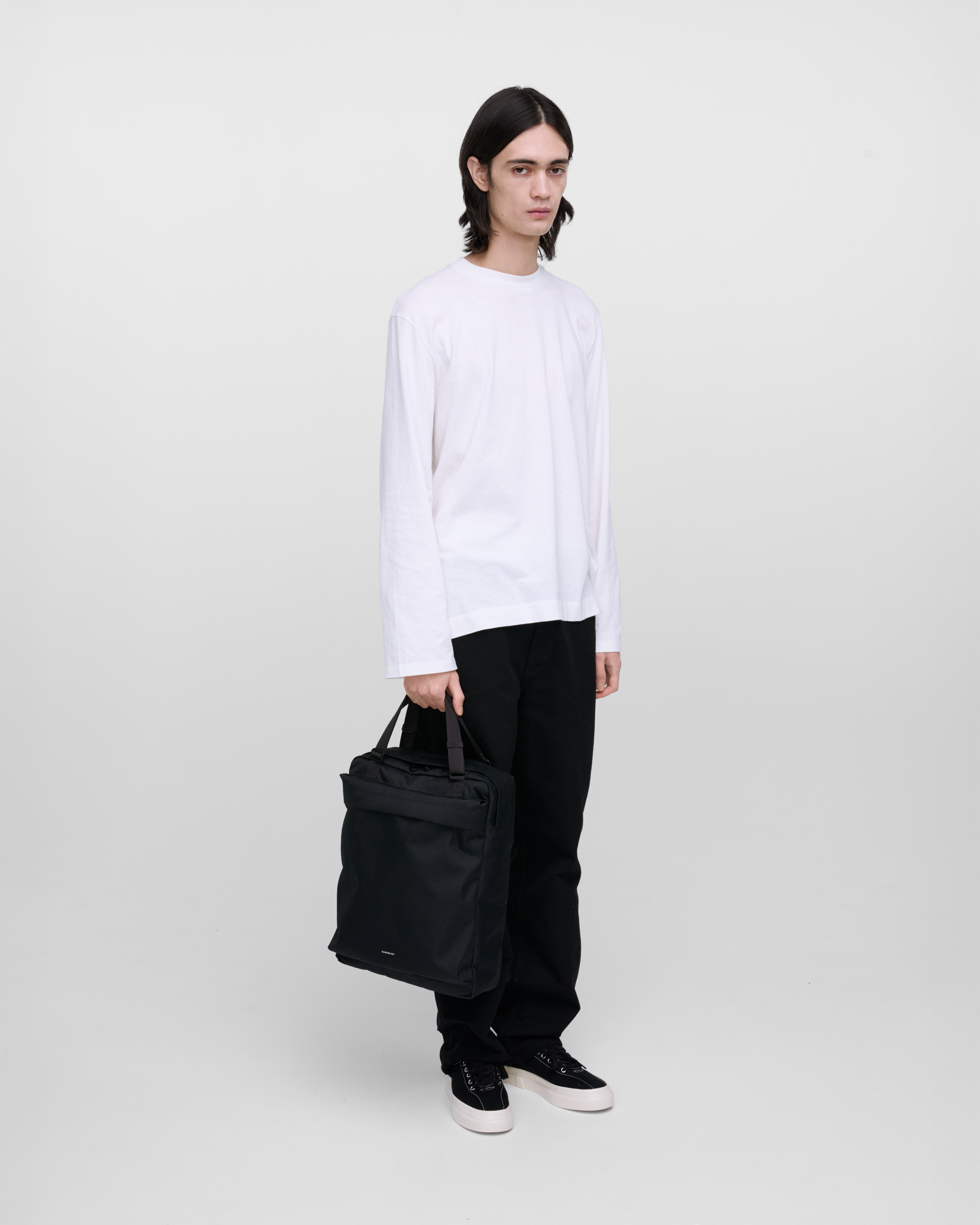 Male model holding Sandqvist Go 2-way Tote Bag, black. Front view, holding the bag in one hand.
