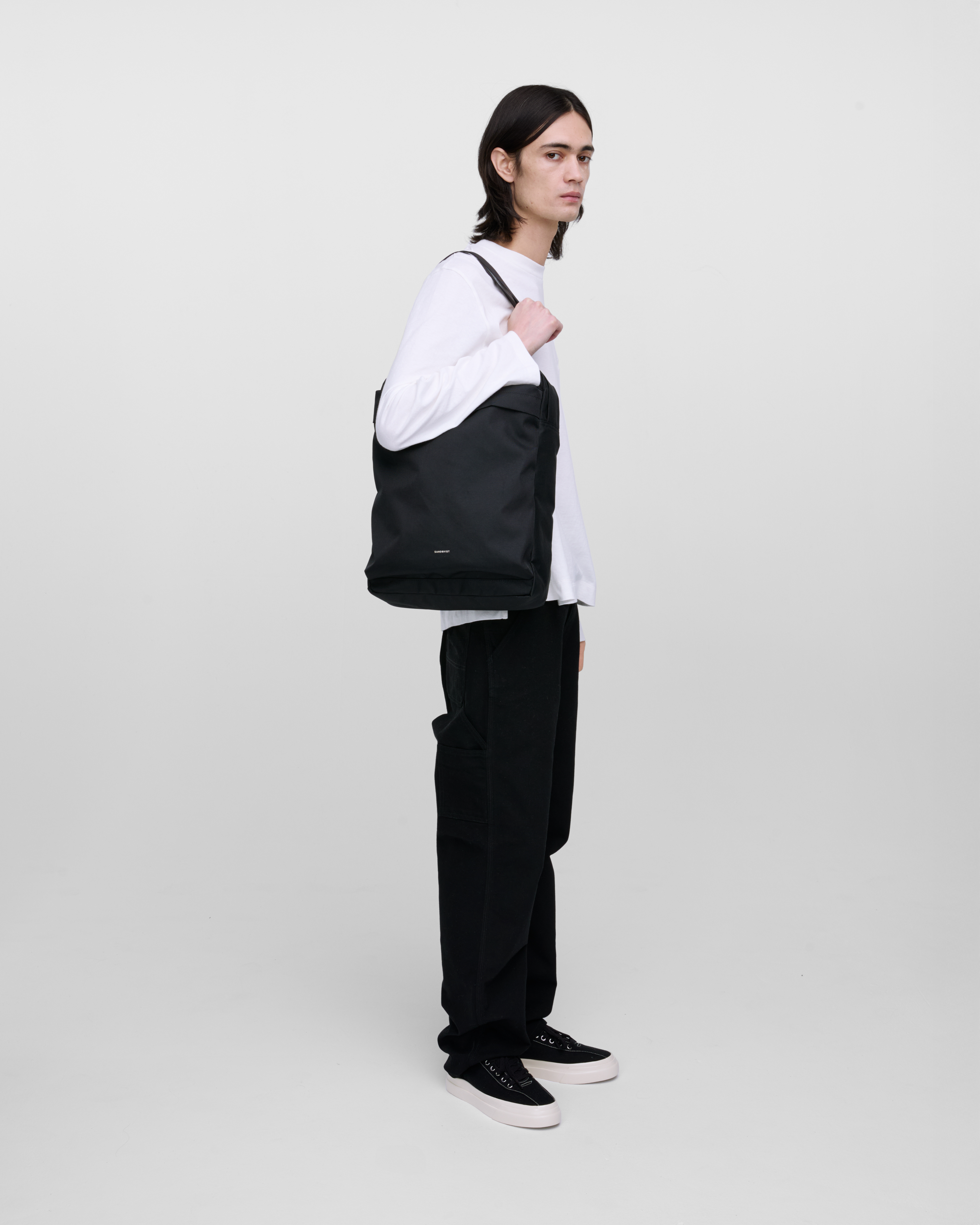 Male model wearing Sandqvist Go 2-way Tote Bag, black. Side view, wearing the bag on one shoulder.