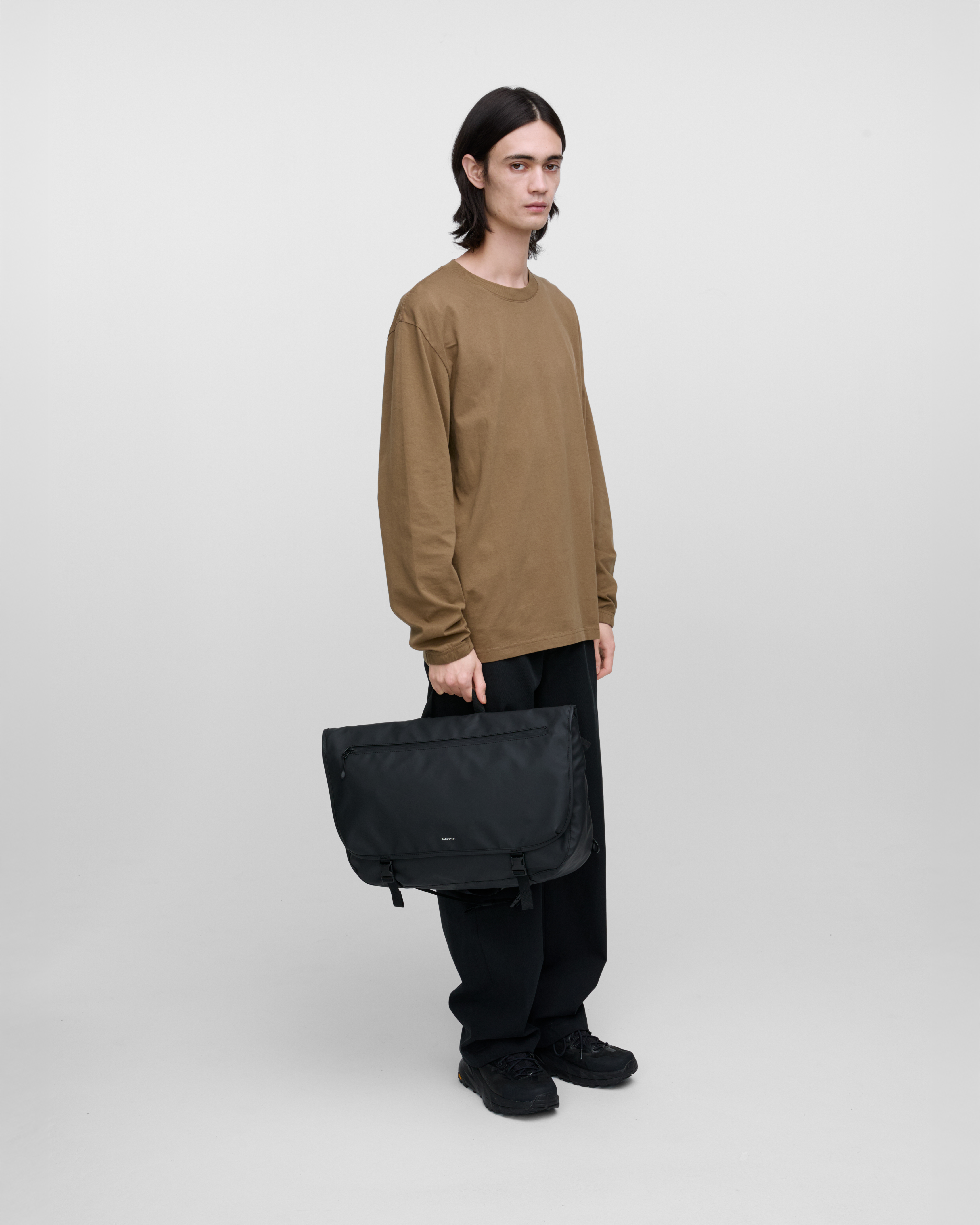 Male model carrying STREAM Messenger Bag Black in the top handle, showing size and shape. 