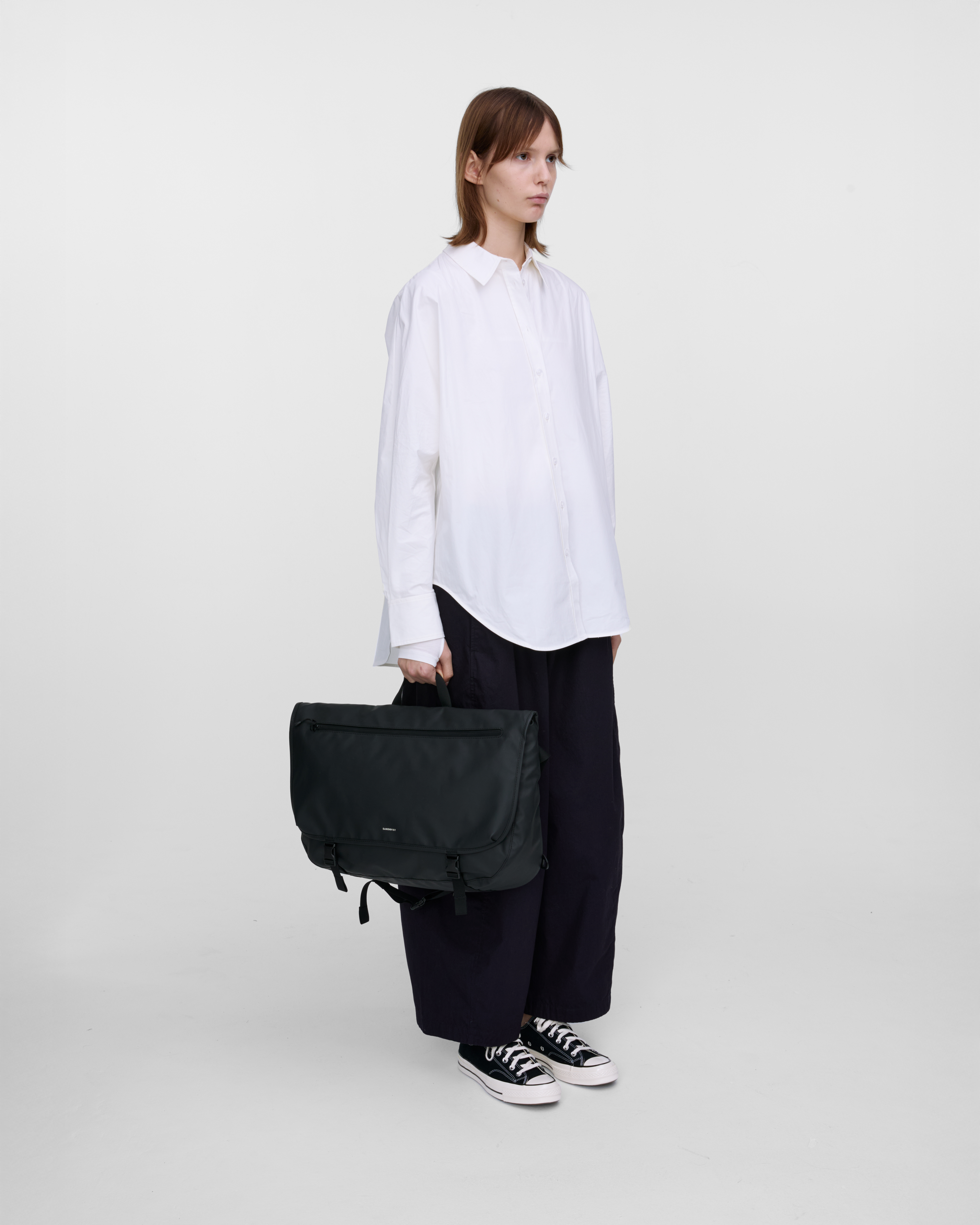 Female model carrying STREAM Messenger Bag Black in the top handle, showing size and shape. 