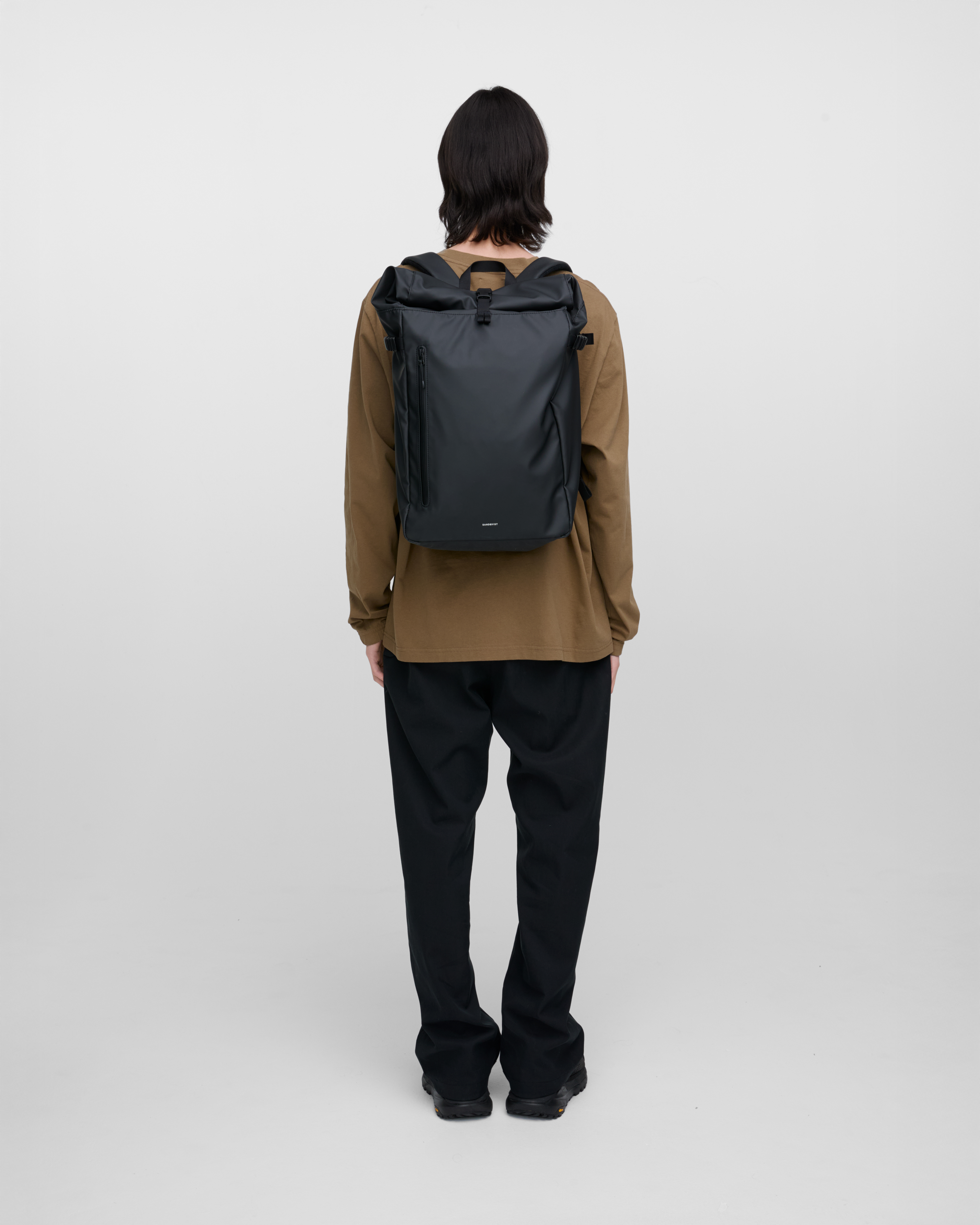 Male model carrying backpack STREAM Slim Rolltop L, in black, back view showing fit and size. 