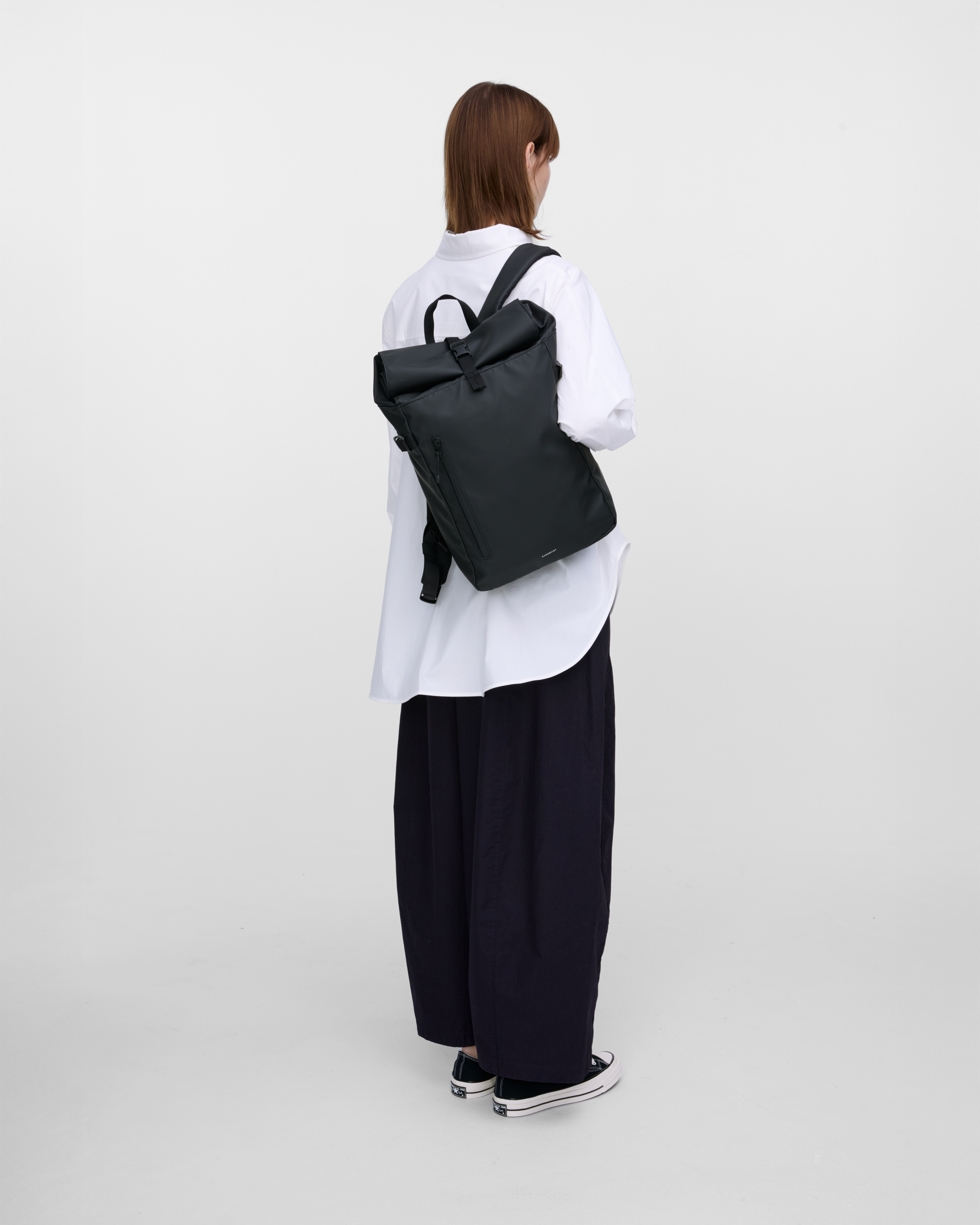 Female model carrying backpack STREAM Slim Rolltop M, Black, hanging over one shoulder, back view showing fit and size. 