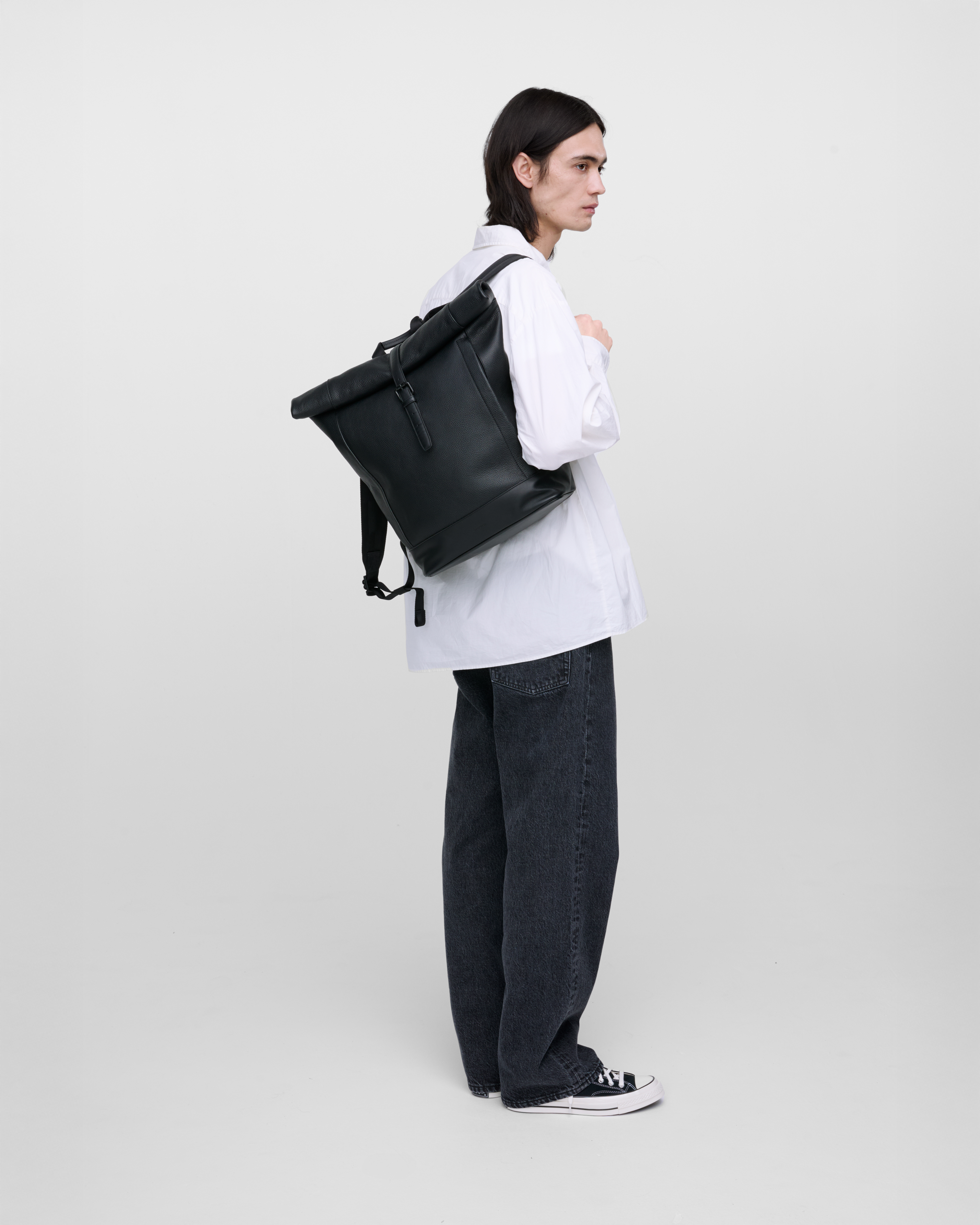Male model carrying the backpack Sandqvist LEATHER Rolltop backpack, Black, hanging over one shoulder, side view showing fit and size.