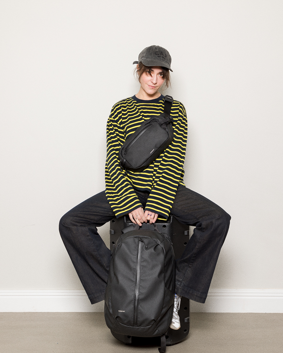 Front view of female model sitting down and wearing a Sandqvist Stream Sling Bag, black and holding a Sandqvist Stream Zipper Backpack 16", black. 