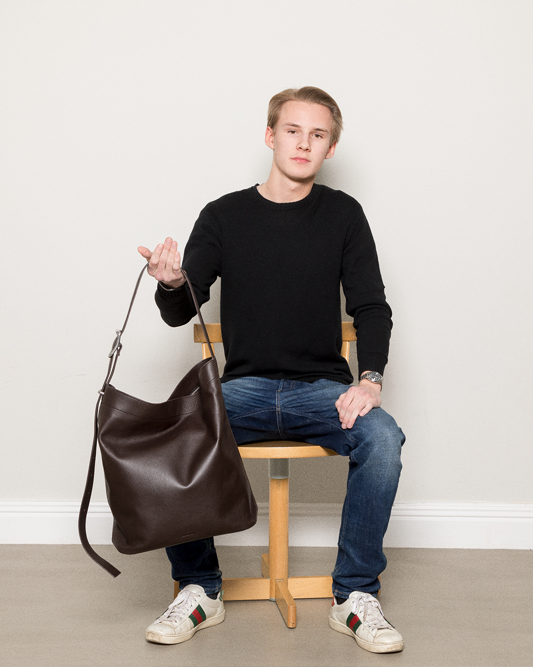 Male model sitting on a chair, holding the Sandqvist Leather Shopper Bag, dark brown. Front view, holding the bag in the handle.