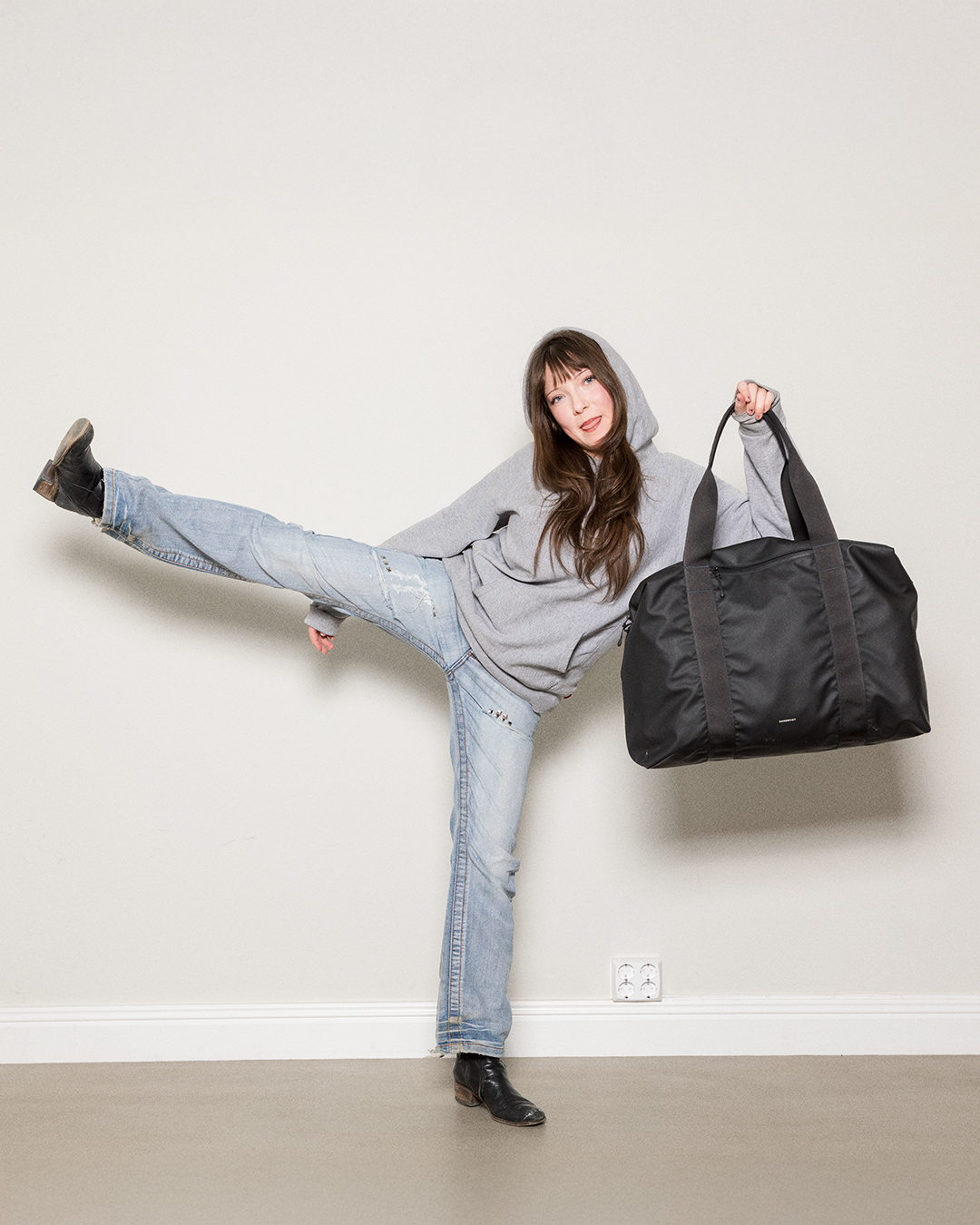 Female model kicking while holding a Sandqvist Stream Weekender Bag, black in one hand. Front view with handles.
