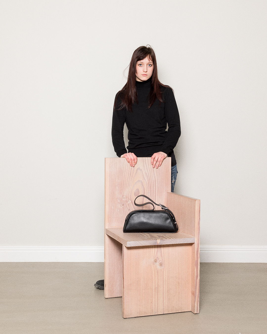 Kvinnlig modell står bredvid en trästol. Sandqvist Leather Baguette Bag ligger på stolen. Framifrån med handtaget synligt.