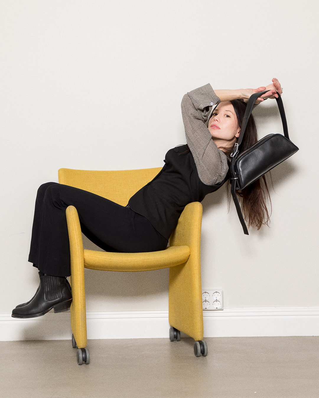 Female model sitting in a yellow chair, holding the Sandqvist Leather Baguette bag, black. 