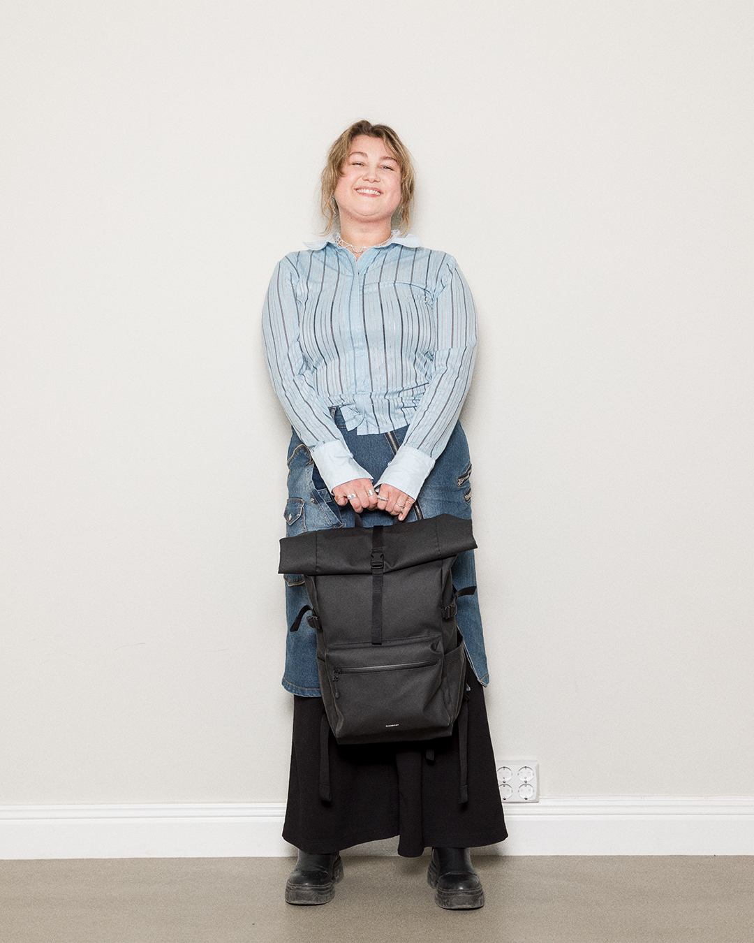 Kvinnlig modell håller Sandqvist Stream Rolltop Ryggsäck L, svart. Framifrån, håller väskan i handtaget. Rolltop och dragkedja fram.