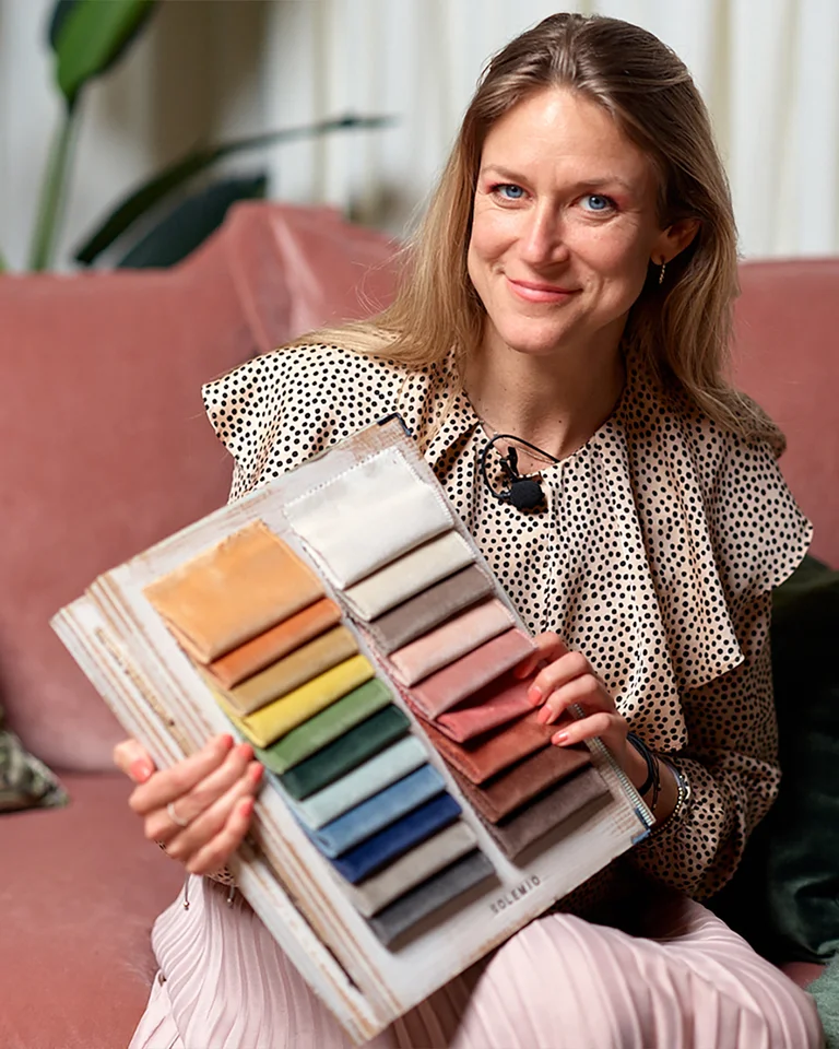 En professionell färgforskare, Sara Garanty, sitter bekvämt i en modern rosa soffa och presenterar en tygkarta med ett urval av färger för anpassningsbara soffklädsel på Sweef. Hennes klädsel och smycken är elegant samordnade med de mjuka tonerna i rummet, som illustrerar konceptet av färgpsykologi i heminredning. Denna bild tjänar som en visuell hjälp för att betona hur valet av soffans färg kan påverka en persons känslor och atmosfären i ett rum.