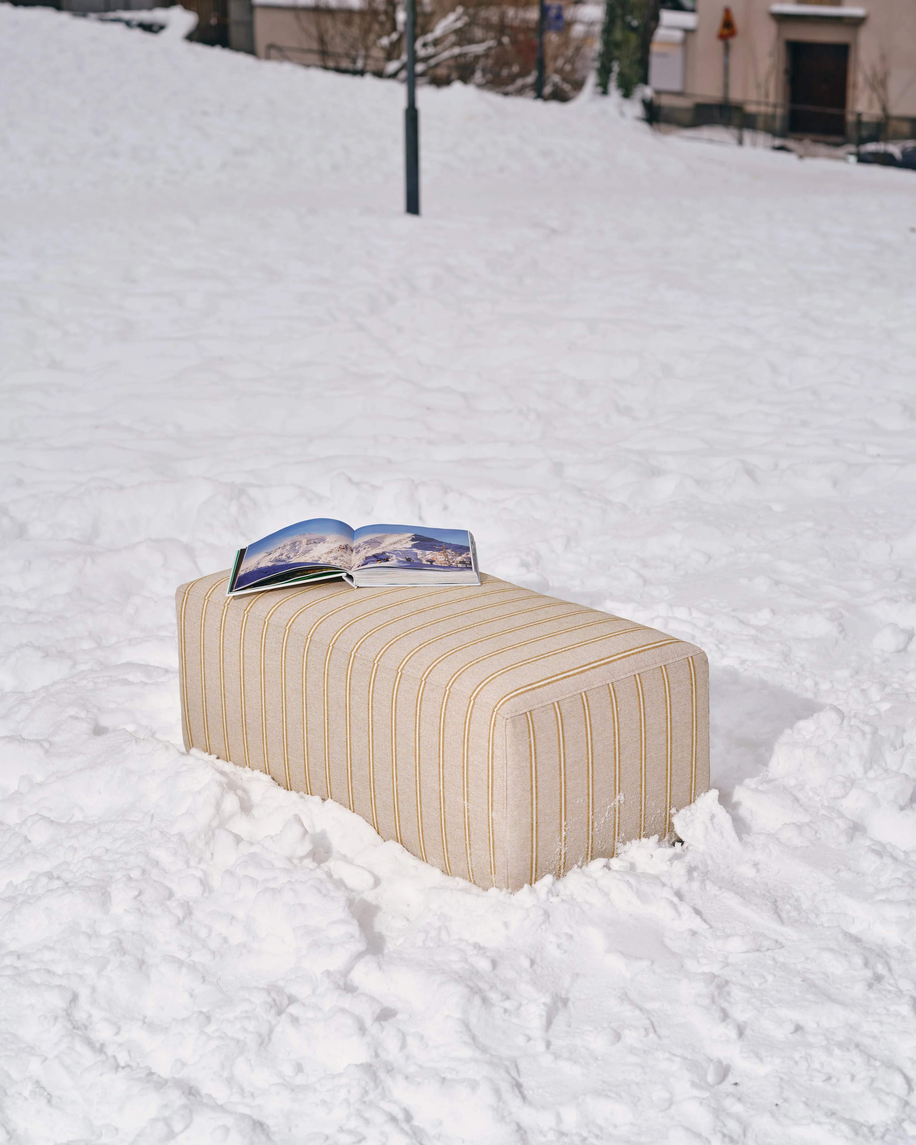 Frittstående fotskammel/puff fra Hajen i stripet gul-beige stoff, plassert i et snødekt landskap.